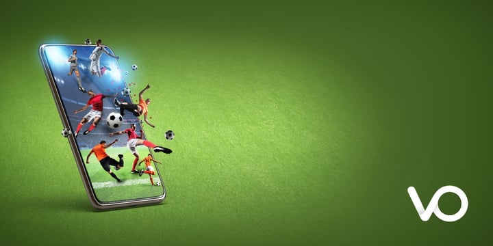 A smartphone on a green surface displays various soccer players and balls bursting out of the screen, symbolizing dynamic sports action. A white VO logo appears in the bottom right corner.