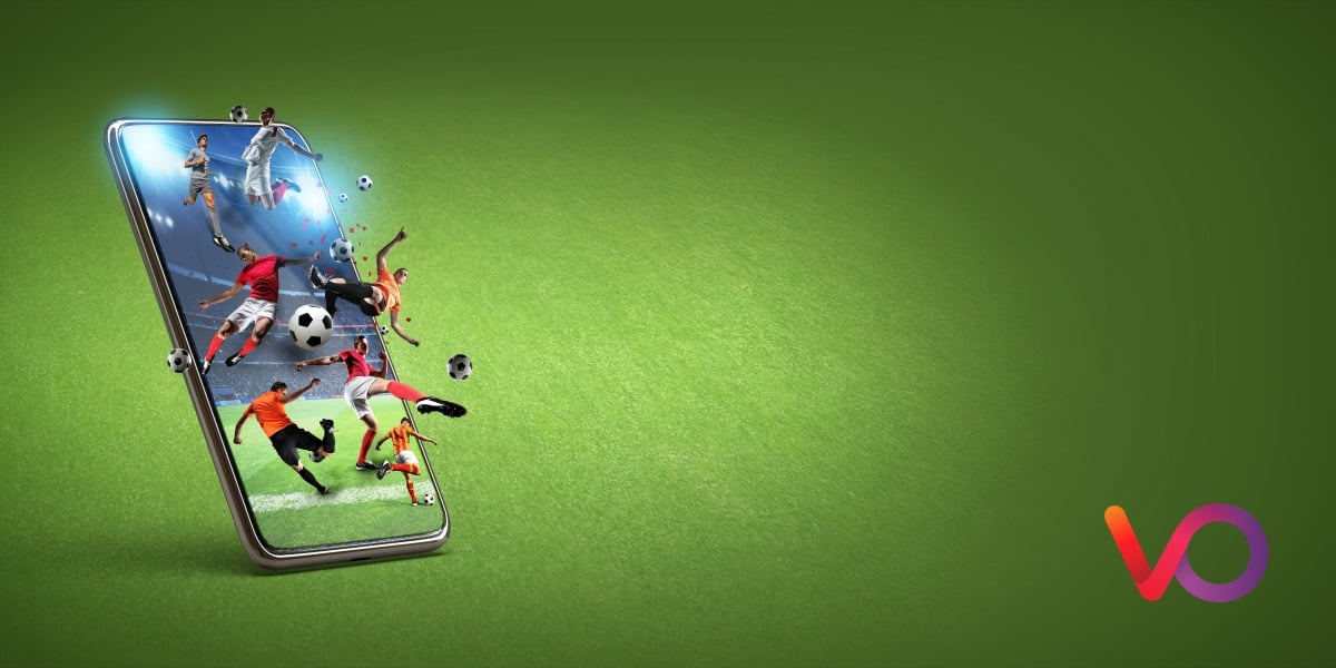A smartphone on a green surface displays various soccer players and balls bursting out of the screen, symbolizing dynamic sports action. A colorful VO logo appears in the bottom right corner.